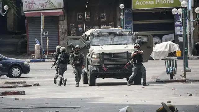جنود إسرائيليون يطلقون الرصاص الحي والغاز المسيل للدموع من أسلحة وخلفهم مركبة عسكرية إسرائيلية. 