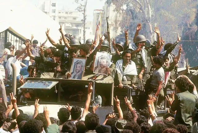 مقاتلون فلسطينيون يظهرون في صورة مؤرخة 22 آب/آب 1982 وهم يرفعون علامات النصر ويحملون صورة زعيمهم ياسر عرفات، رئيس منظمة التحرير الفلسطينية، فوق شاحنة عسكرية في بيروت، أثناء توجههم إلى تونس