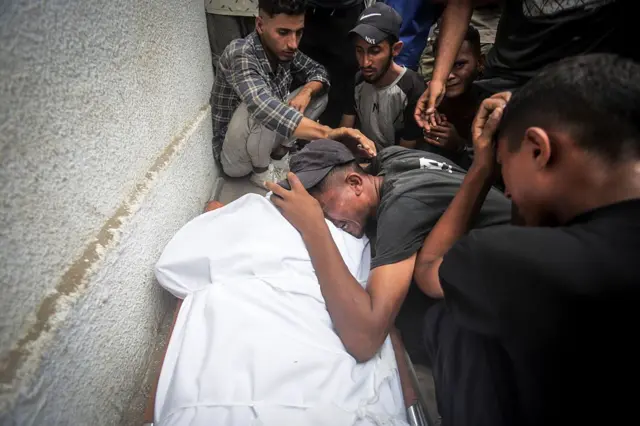 أقارب لفلسطينيين لقوا حتفهم، نتيجة للهجمات الإسرائيلية على أجزاء مختلفة من مدينة غزة، ينعون أقاربهم الموتى في مستشفى الشفاء خلال إجراءات جنازتهم في 27 آب/آب 2025، وتظهر في الصورة جثة مغطاة بالكفن الأبيض، بينما يبكي شخص بجوارها محاطاً بأشخاص أخرين يواسوه.