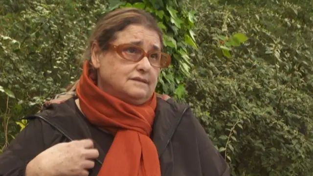 امرأة ترتدي نظارة بنية اللون ومعطفًا بنيًا ووشاحًا برتقاليًا، تنظر إلى جانب الكاميرا وتقف أمام سياج.