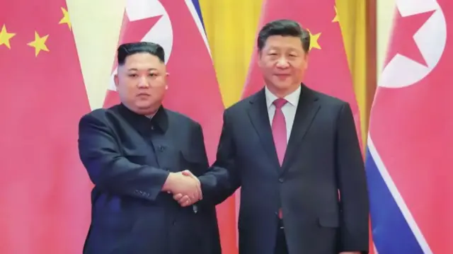 كيم جونغ أون وشي جينبينغ يقفان أمام أعلام، ويتصافحان مع تعبيرات جادة على وجوههما
